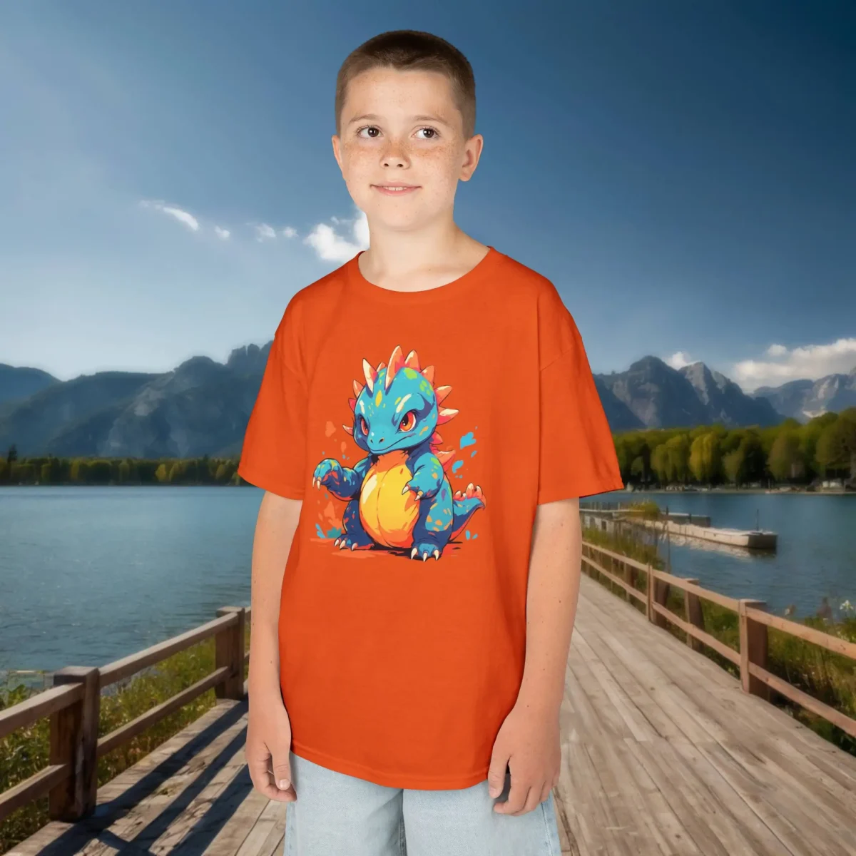 Boy modeling an orange kids T-shirt with a vibrant baby Therizinosaurus print, standing on a dock with lake and mountain views.