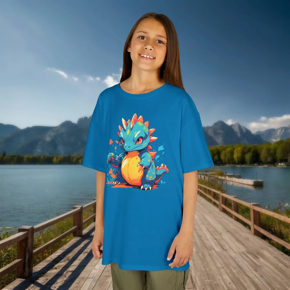 Girl wearing a sapphire blue T-shirt with a bold baby Therizinosaurus design, standing outdoors by a docked lake with mountains in the background.