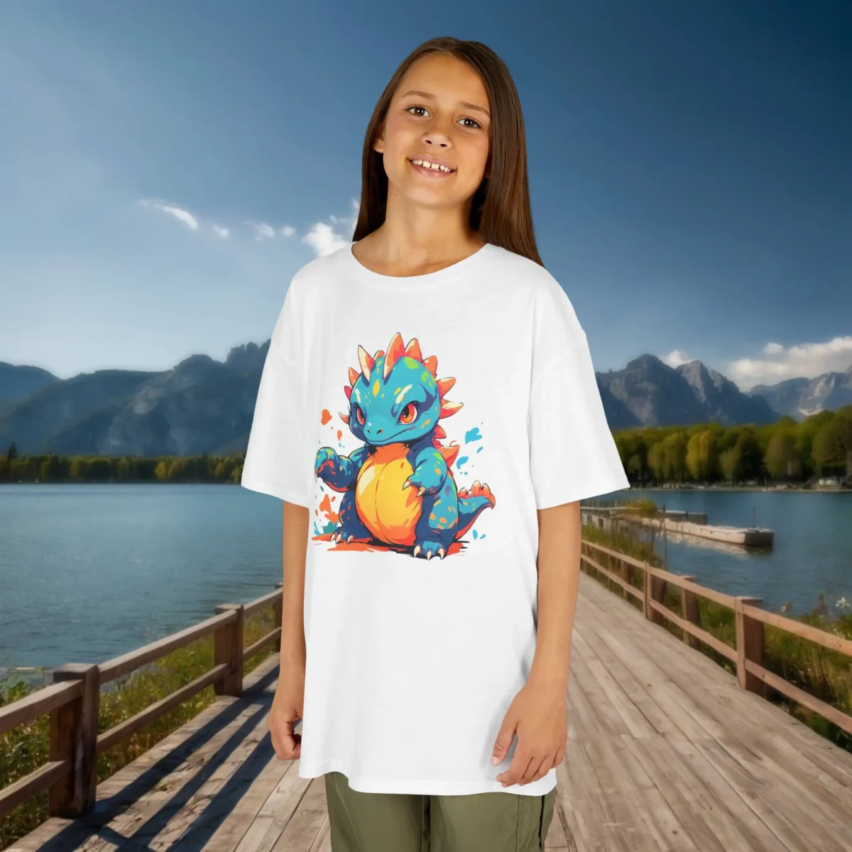 Girl wearing a white kids T-shirt featuring a fierce and colorful baby Therizinosaurus illustration, standing on a dock by a lake with mountains in the background.