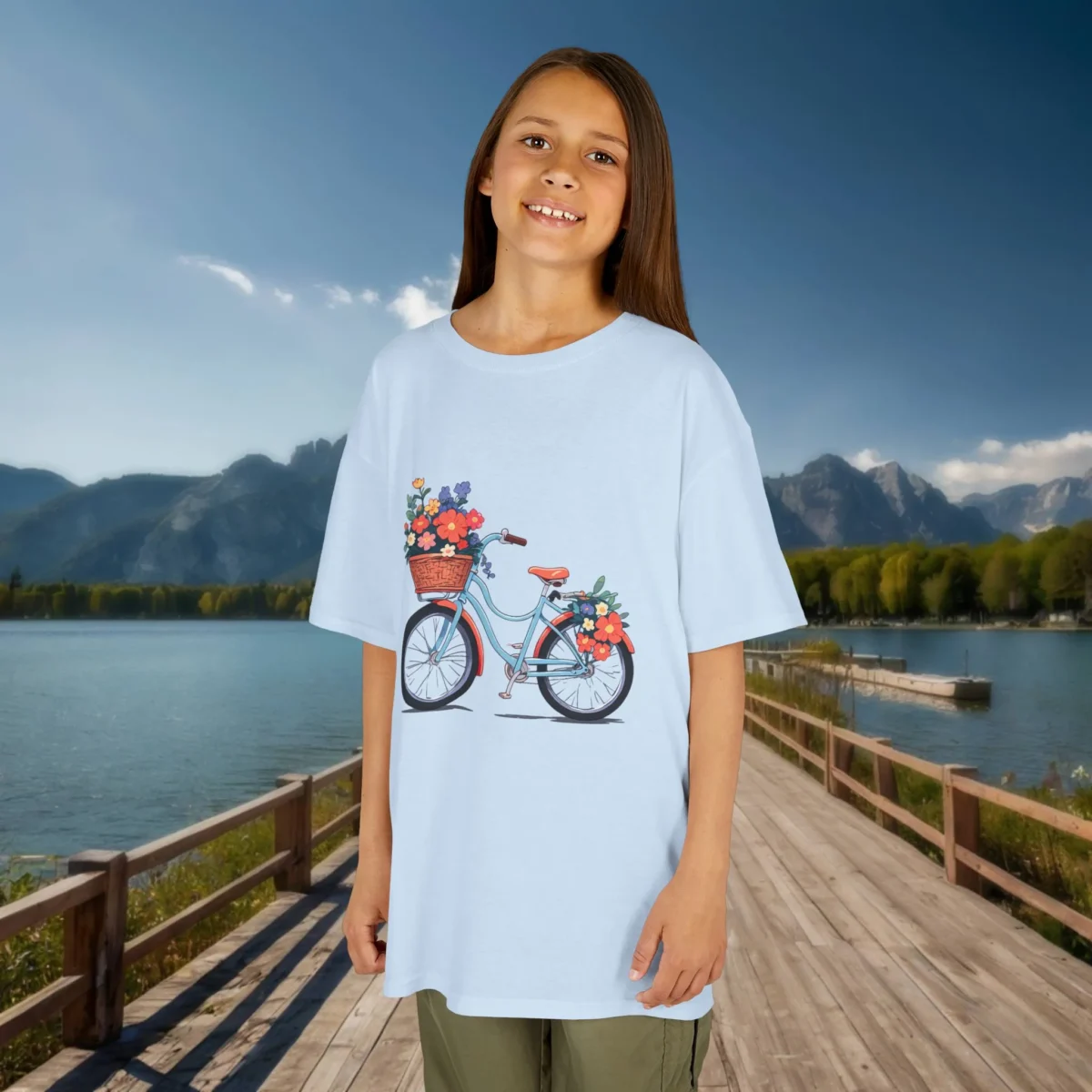 Smiling girl wearing a light blue kids t-shirt with a colorful bicycle and flower print design, standing on a lakeside boardwalk.