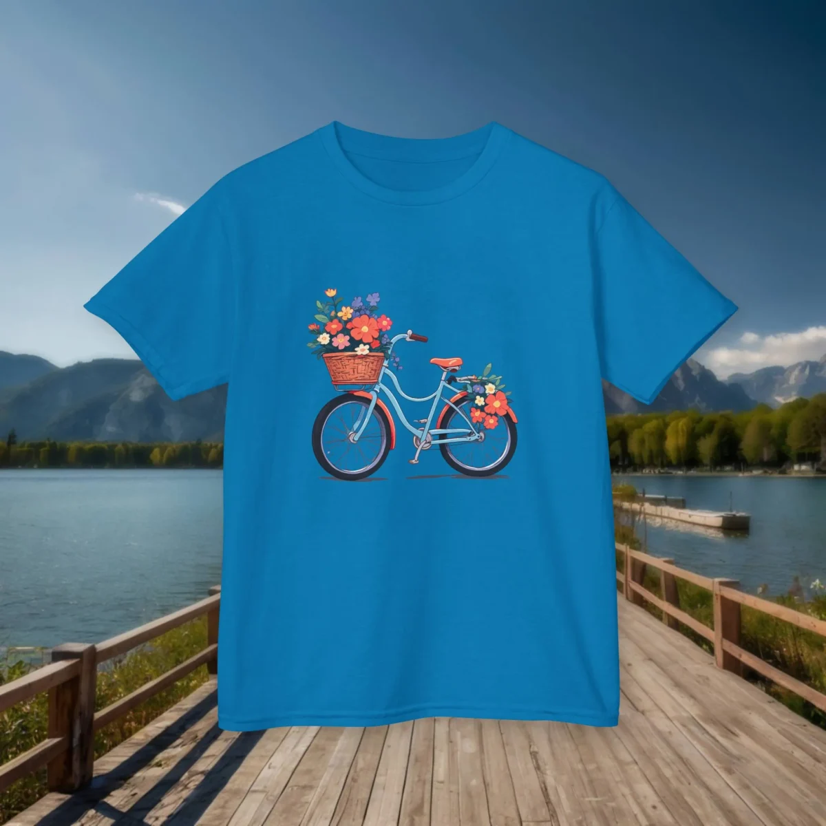 Flat lay of a royal blue kids t-shirt with a colorful bicycle and flower graphic, displayed on a lakeside wooden boardwalk.