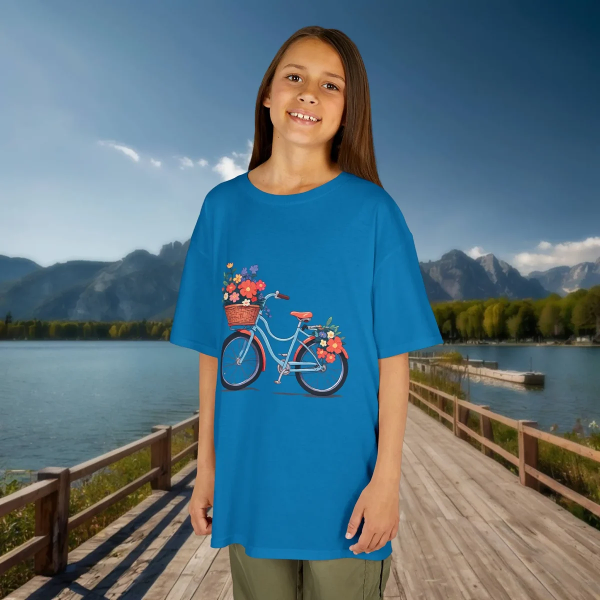 Girl wearing a royal blue t-shirt with a floral bicycle graphic, smiling on a wooden walkway beside a lake and mountains.