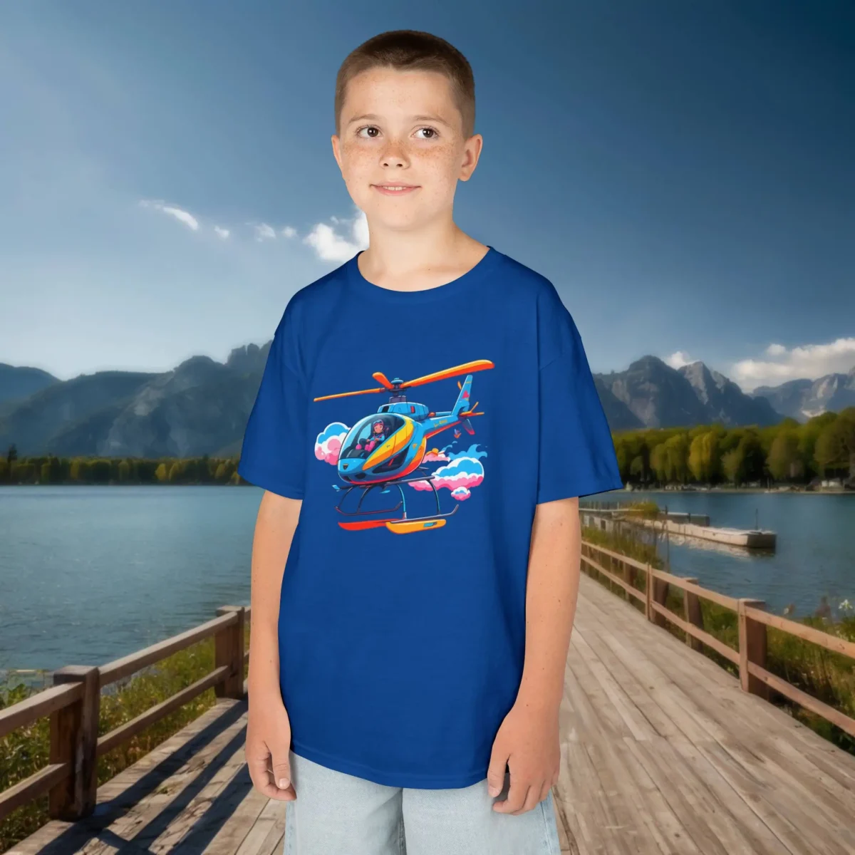 Boy wearing a royal blue kids t-shirt with a colorful cartoon helicopter flying above clouds, posing on a dock by a mountain lake.
