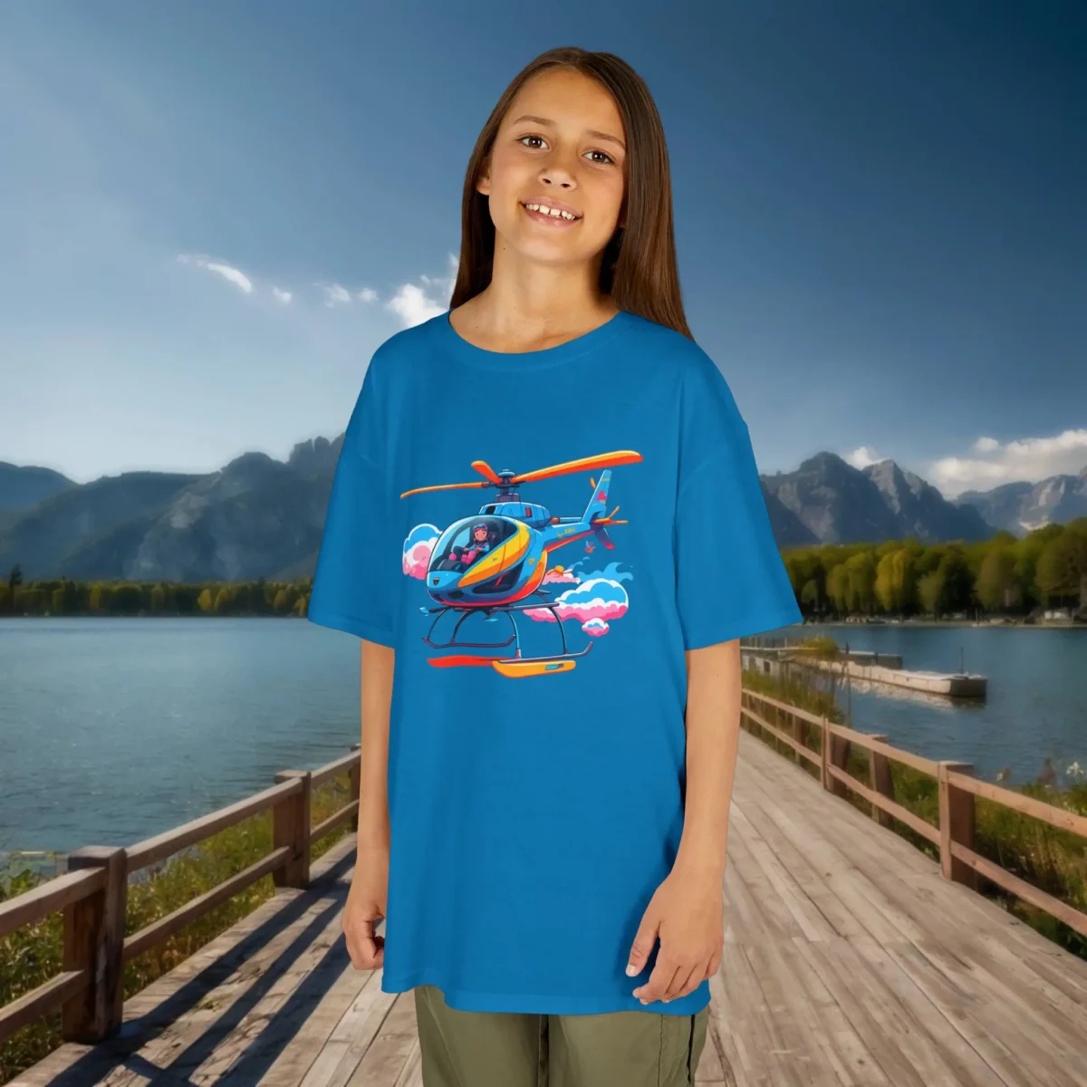 Smiling girl wearing a sapphire blue kids t-shirt with a colorful cartoon helicopter flying through clouds, standing on a boardwalk by a scenic lake.
