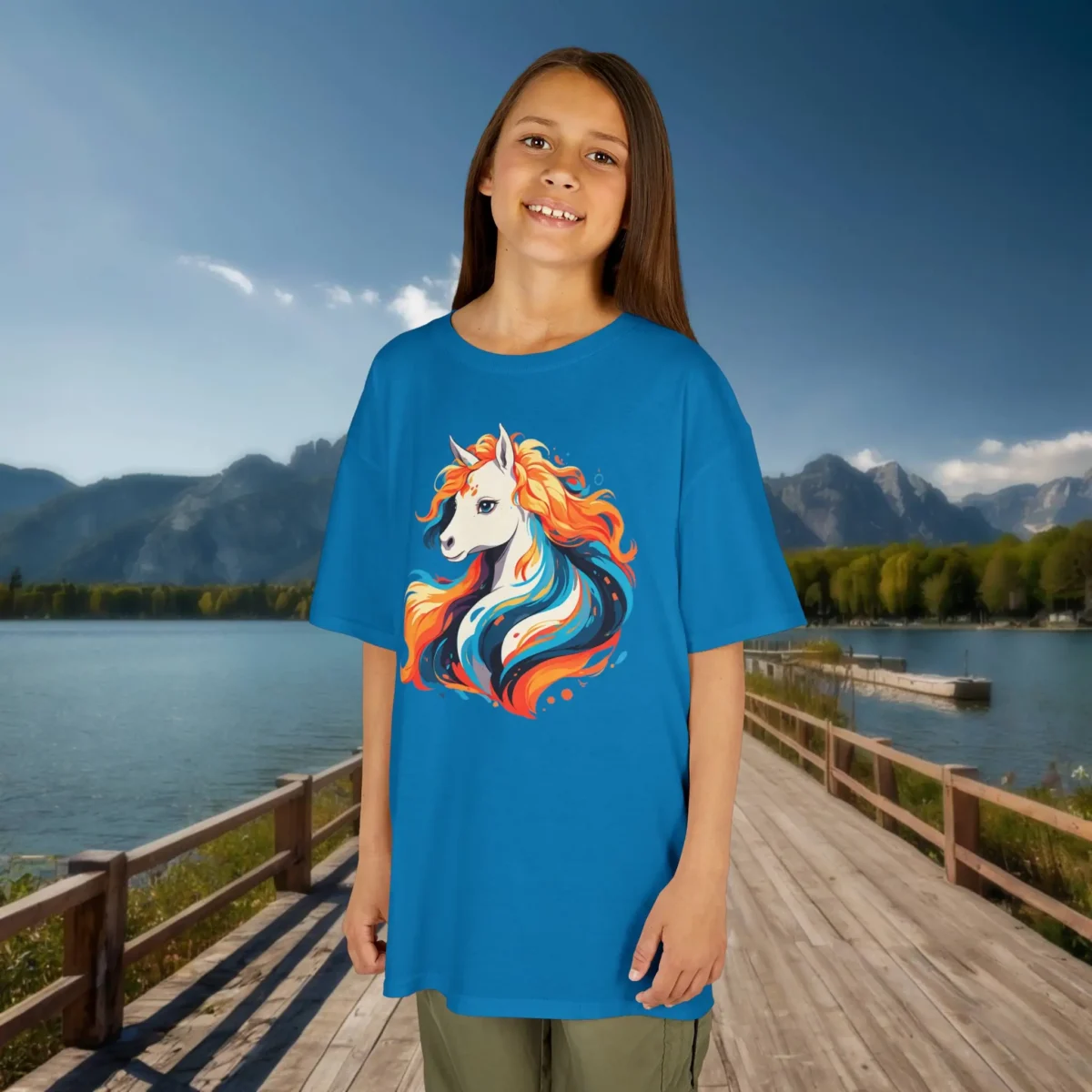 Girl wearing a sapphire blue t-shirt featuring a white unicorn with a fiery, colorful mane, posing on a scenic boardwalk by a mountain lake.