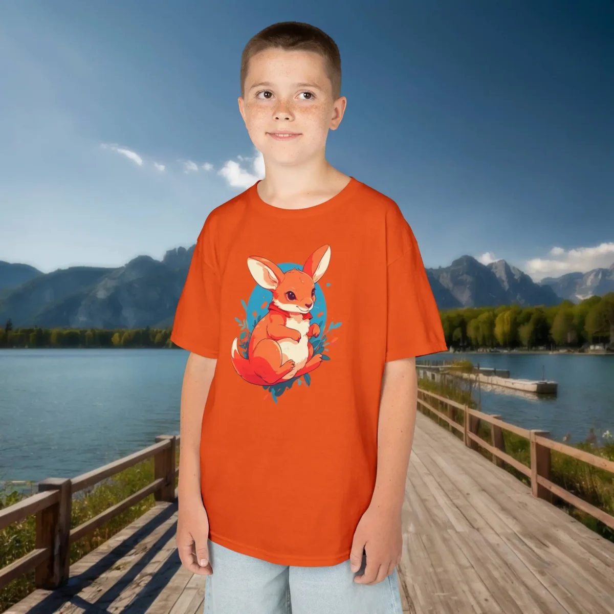 Boy wearing an orange kids t-shirt with a cute cartoon kangaroo sitting on grass with a blue background, standing by a mountain lake.