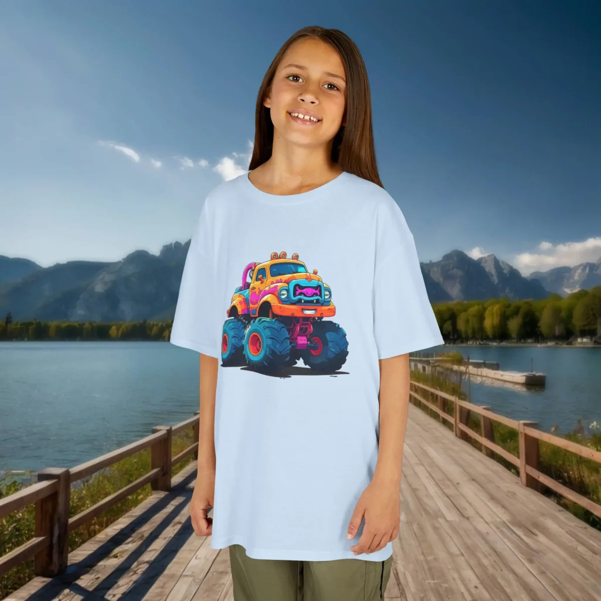 Girl wearing a light blue kids t-shirt featuring a colorful cartoon monster truck with big wheels and bright details, standing on a wooden dock by a lake.