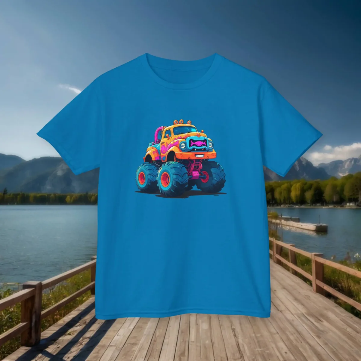 Flat lay of a sapphire blue kids t-shirt with a colorful cartoon monster truck graphic, placed on a dock with a lake and mountains in the background.