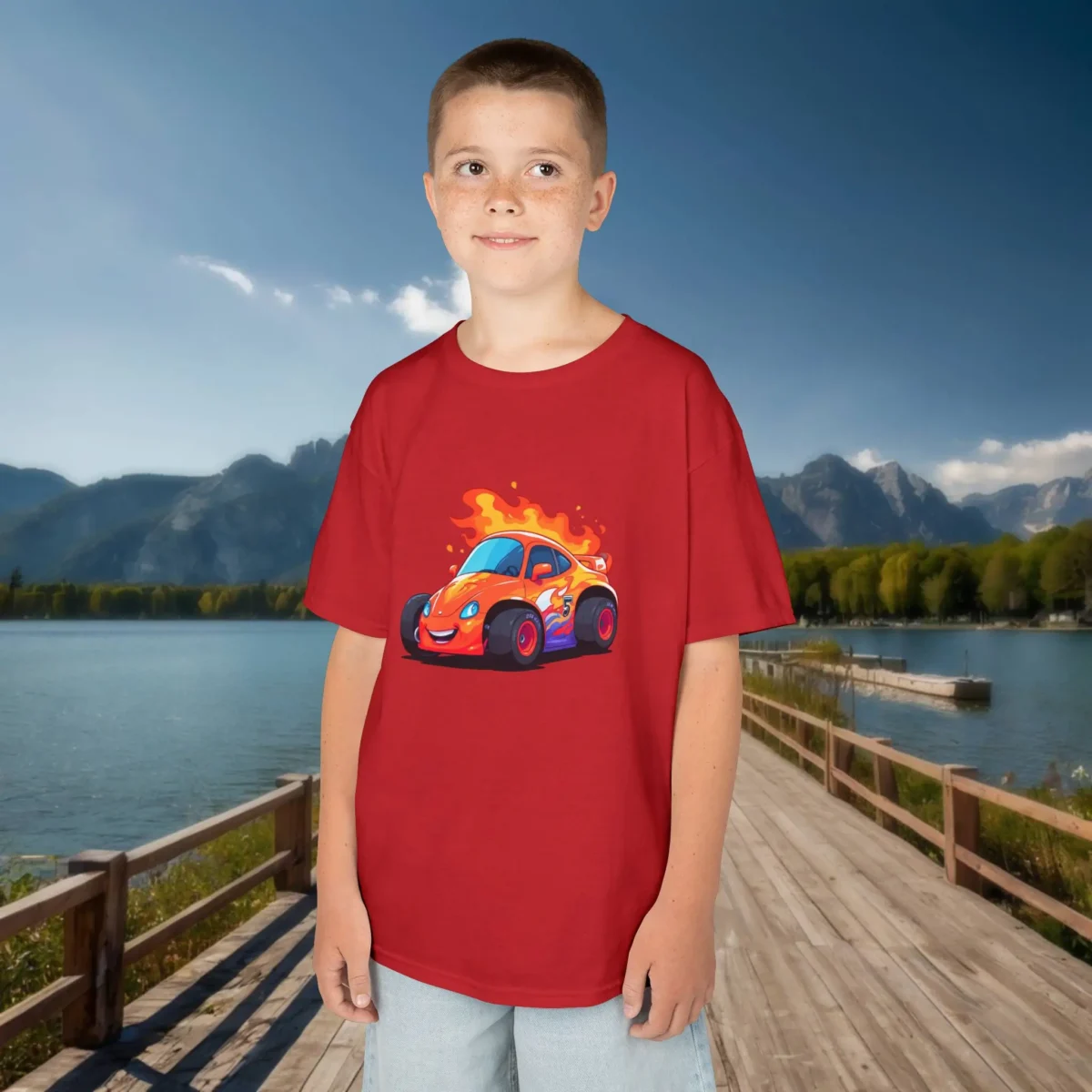 Boy wearing a red kids t-shirt with a cartoon race car engulfed in flames, standing on a dock by a mountain lake.