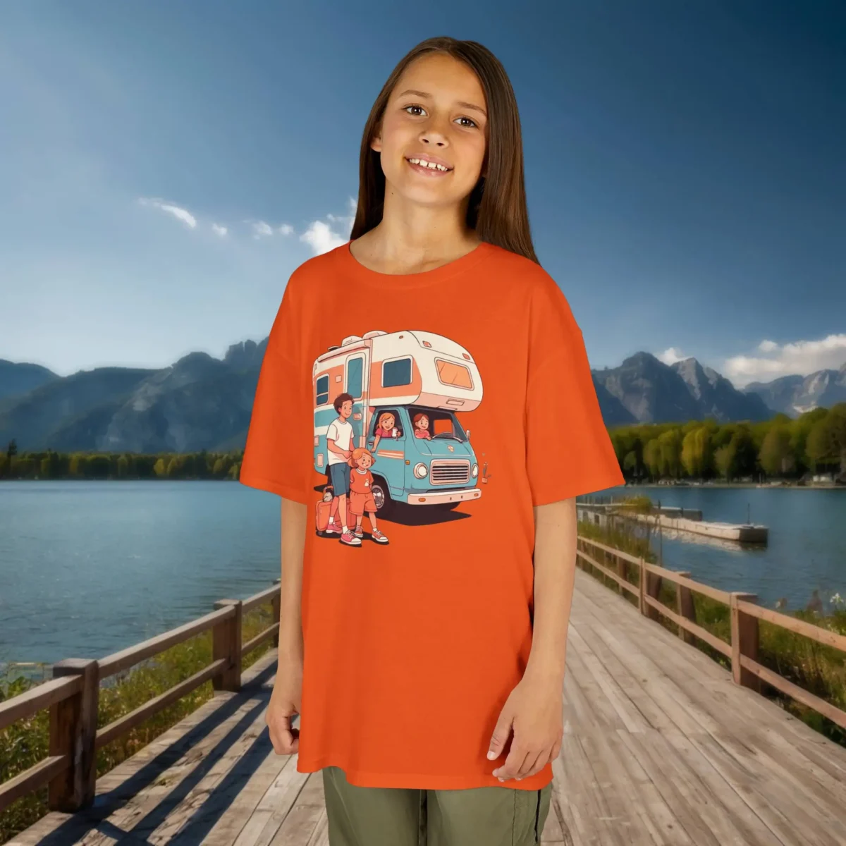 Girl wearing an orange kids t-shirt featuring a happy cartoon family and RV camper, with a lake and mountains in the background.