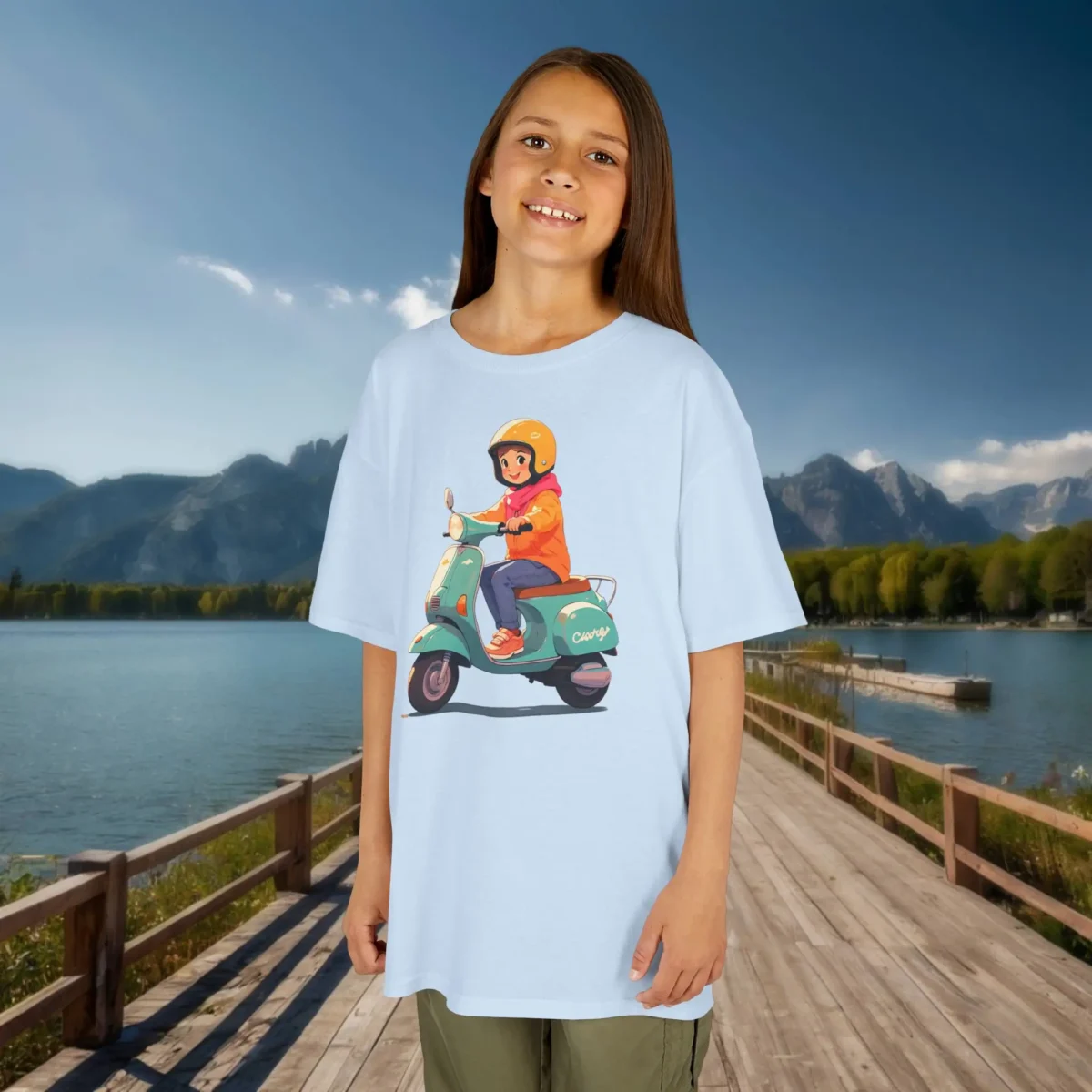 Girl wearing a light blue kids t-shirt with a cartoon of a girl riding a scooter, in a scenic lakeside setting.