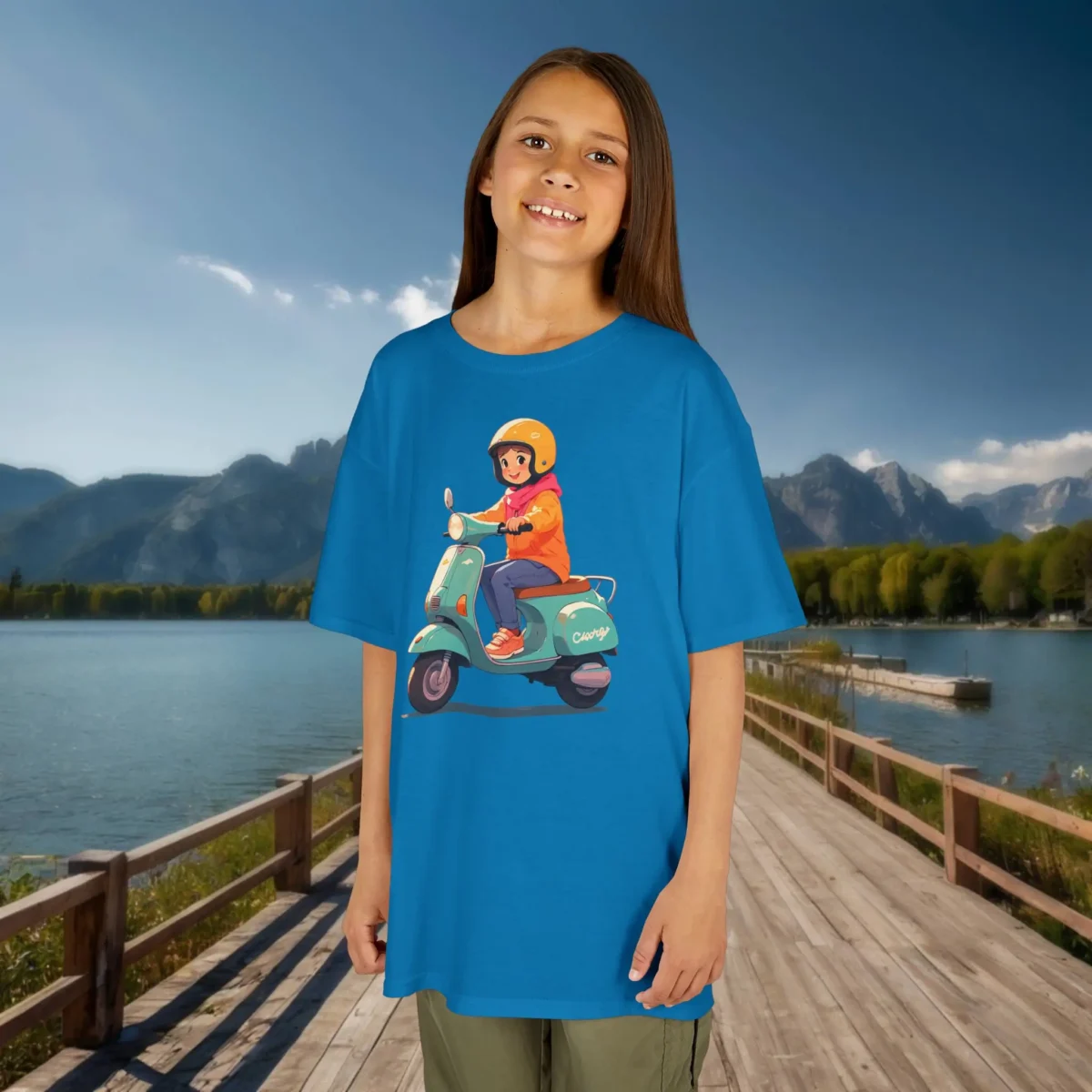 Girl wearing a sapphire blue kids t-shirt with a cartoon girl on a mint-green scooter, standing on a wooden dock by a lake.