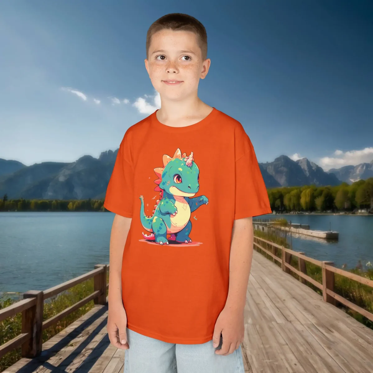 Boy wearing an orange t-shirt with a colorful cartoon baby Iguanodon that has a small horn and cheerful expression, standing near a mountain lake.