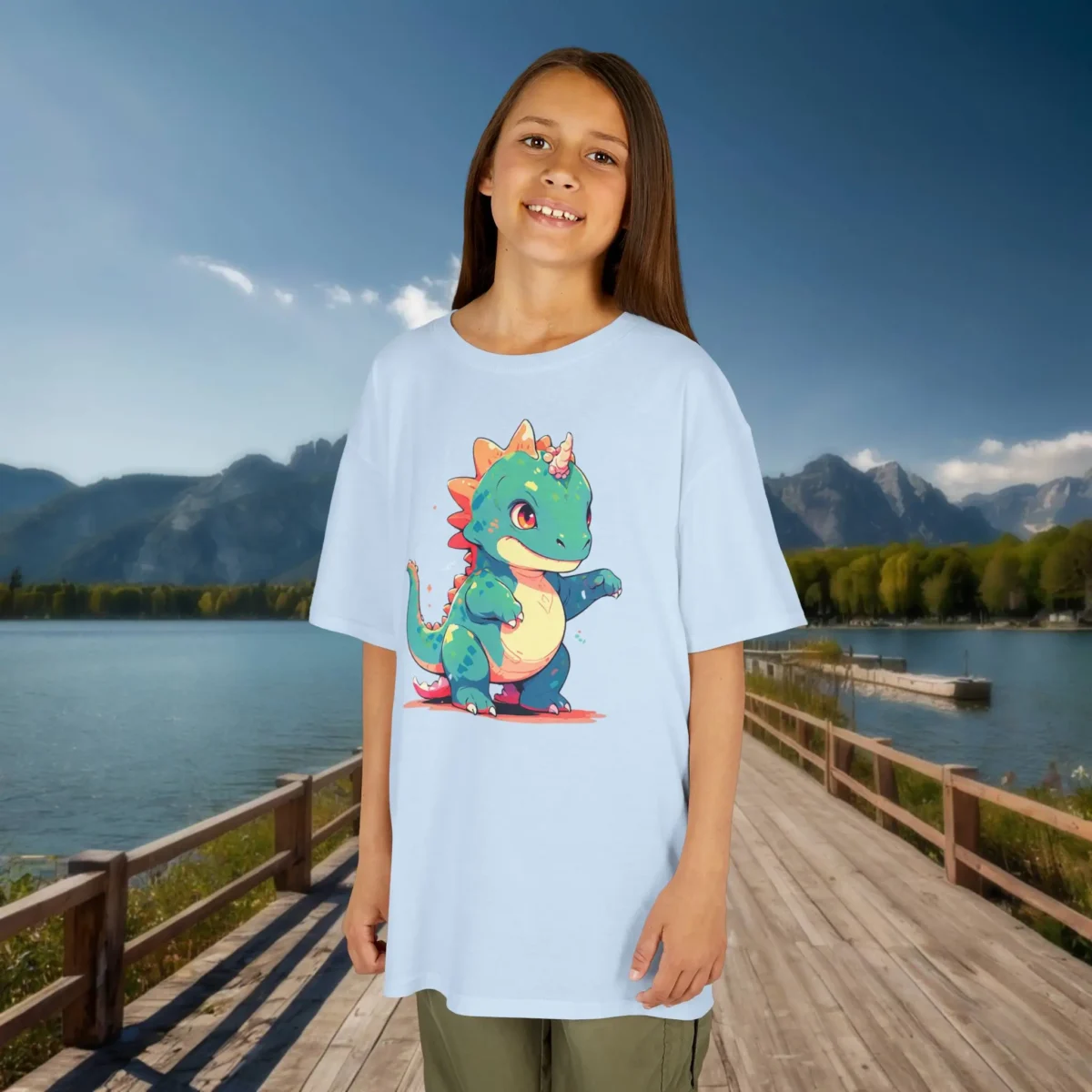 Girl wearing a sky blue t-shirt with a cute cartoon baby Iguanodon featuring a colorful horn and tail, standing on a dock by the lake.