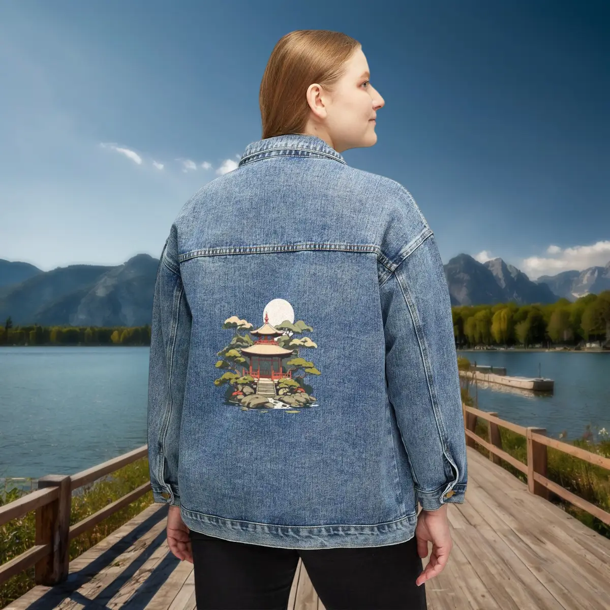 Women’s Denim Jacket with Japanese Temple Print – Lifestyle View Woman wearing a blue denim jacket with a Japanese temple illustration on the back, standing on a wooden boardwalk near a lake with mountains in the background