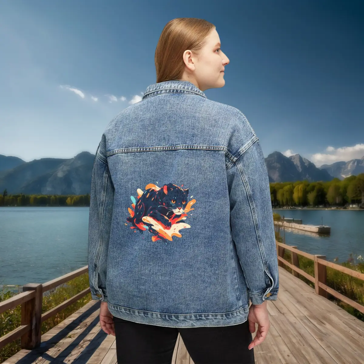 Women's Denim Jacket with Jaguar Print – Back View Woman wearing a denim jacket with colorful jaguar illustration on the back, standing on a wooden deck near a lake.