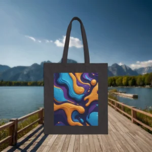 Flat black cotton tote bag with a vivid, abstract oil spill design placed on a wooden pier with mountains and a lake in the background.