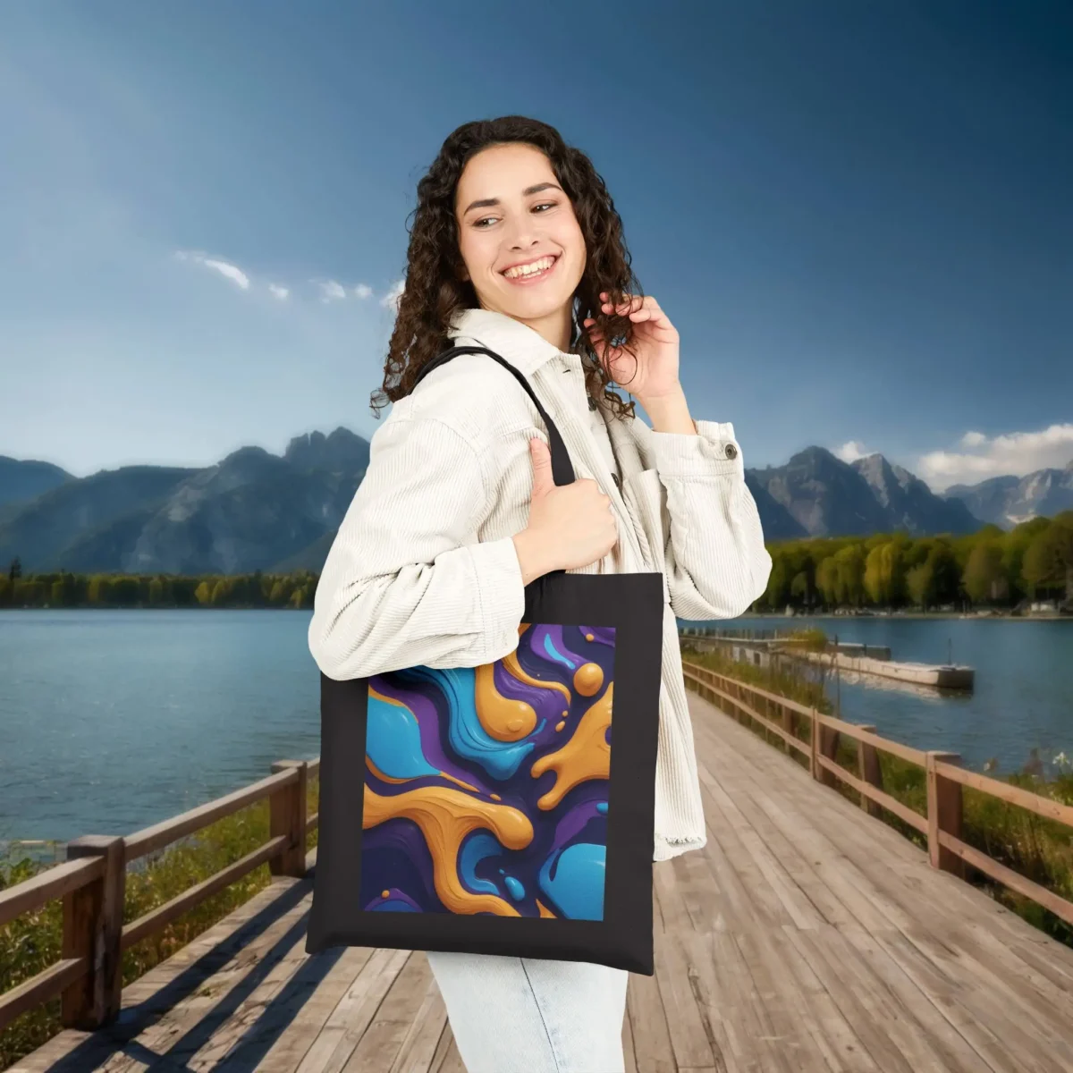 Woman carrying a black cotton tote bag featuring a bold and colorful abstract oil spill pattern, standing on a wooden dock near a lake.